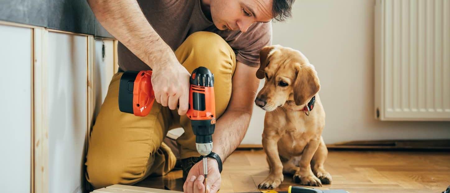 Man som utför renovering, potentiell mottagare av ett renoveringslån, borrar i en planka med sin hund bredvid.